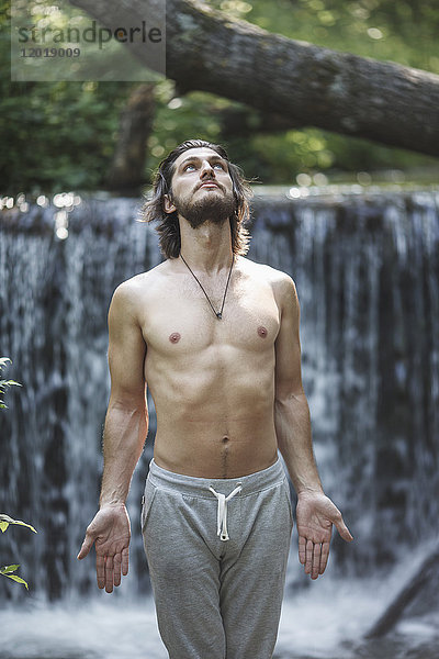 Hemdenloser junger Mann schaut auf  während er am Wasserfall im Wald steht.