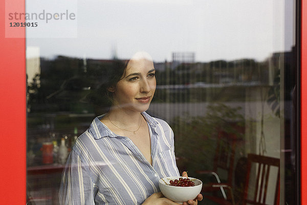 Nachdenkliche junge Frau hält Schale mit roten Johannisbeeren beim Blick durchs Fenster