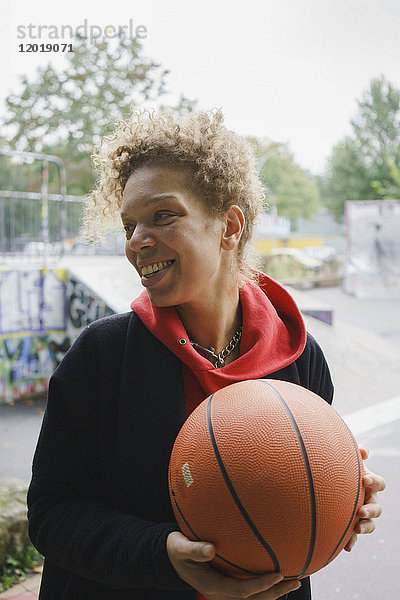 Lächelnde erwachsene Frau hält Basketball während sie im Park steht.