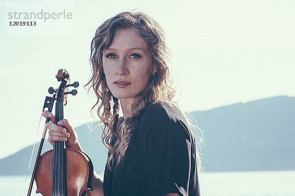 Porträt einer selbstbewussten jungen Frau  die an einem sonnigen Tag Geige gegen den klaren Himmel hält.