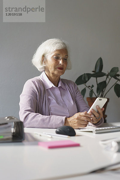 Lächelnde Seniorin schaut auf das Handy  während sie im Büro an der Wand sitzt.