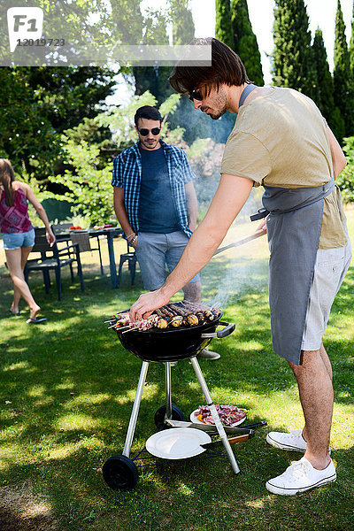 Gut aussehender junger Mann  der bei einer Grillparty im Garten während des Sommerurlaubs Fleisch zubereitet.