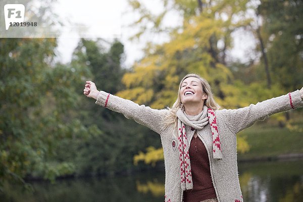 Porträt einer jungen blonden Frau  die an einem See im Herbst mit den Armen in die Luft lächelt.
