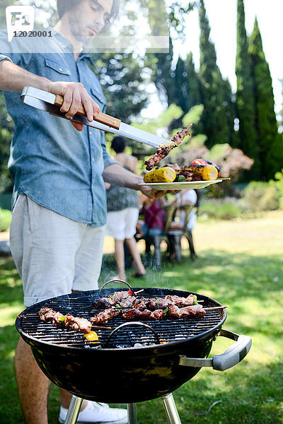 Gut aussehender junger Mann  der bei einer Grillparty im Garten während des Sommerurlaubs Fleisch zubereitet.