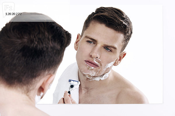 Oben-ohne-Mann  der in den Spiegel schaut  mit Rasierschaum auf seiner Gesichtshälfte  mit Rasiermesser in den Händen  rasierend