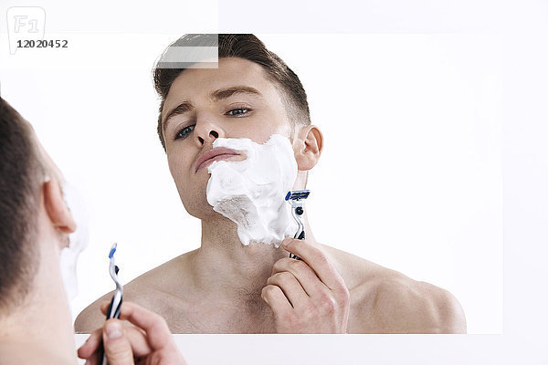 Oben-ohne-Mann  der in den Spiegel schaut  mit Rasierschaum auf seiner Gesichtshälfte  mit Rasiermesser in den Händen
