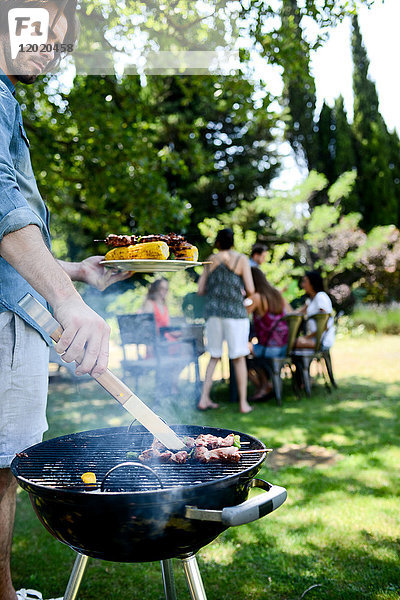 Gut aussehender junger Mann  der bei einer Grillparty im Garten während des Sommerurlaubs Fleisch zubereitet.