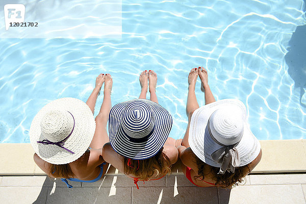 Drei schöne junge Frau mit Badeanzug und Sonnenhut sitzen am Rande des Pools der Anlage Schwimmbad während des Sommerurlaubs.