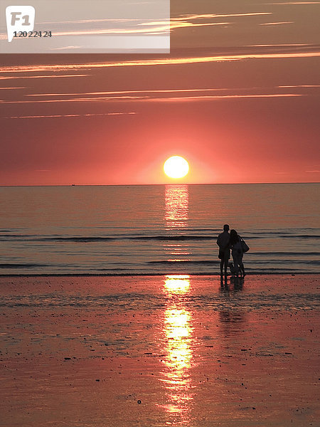 Normandie  Paar mit Hund im Hintergrund betrachtet den Sonnenuntergang an einem Strand