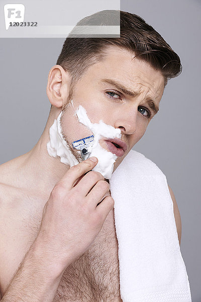 Oben-ohne-Mann  Handtuch auf der Schulter  mit Rasierschaum auf der Gesichtshälfte  mit Rasiermesser in den Händen  ernst  rasierend  offener Mund