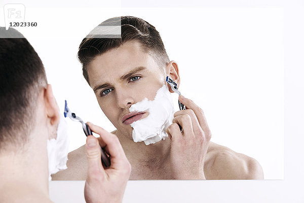 Oben-ohne-Mann  in den Spiegel schauend  rasierend  mit Rasierapparat in der Hand  Rasierschaum auf der Hälfte seines Gesichts