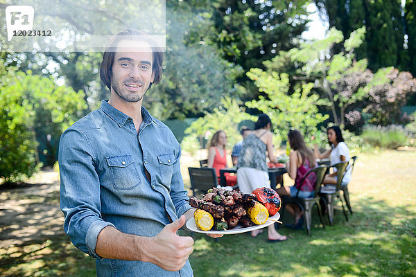 Hübscher junger Mann zeigt gegrilltes Fleisch in Grillparty im Freien im Garten während des Sommerurlaubs.