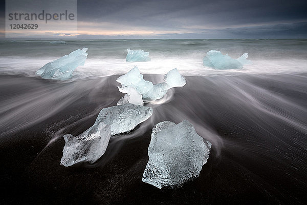Gläserne Eisstücke auf vulkanischem schwarzem Sandstrand mit verschwommenen Wellen  in der Nähe der Lagune Jokulsarlon  Südisland  Polarregionen