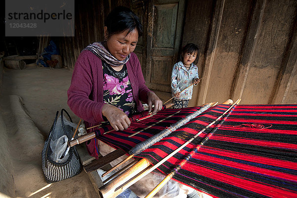 Frau beim Weben des traditionellen roten Naga-Schals  daneben ihre kleine Tochter  auf der Veranda des Familienhauses  Nagaland  Indien  Asien