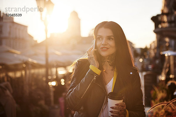 Junge Frau geht im Freien  hält Kaffeetasse  benutzt Smartphone  Tätowierungen an Händen und Hals