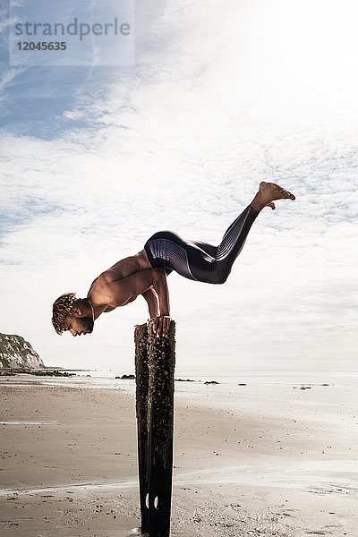 Junger Mann trainiert  macht Handstand mit erhobenen Beinen auf hölzernen Strandpfosten