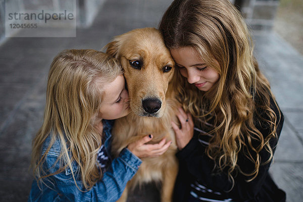 Mädchen und ihre Schwester sitzen auf der Terrasse und streicheln ihren Golden Retriever.