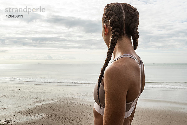 Junge weibliche Läuferin mit Haarzöpfen  die vom Strand aufs Meer blickt