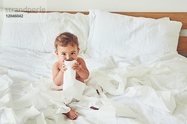 Kleinkind sitzt auf dem Bett und hält aufgetrennte Toilettenpapierrolle