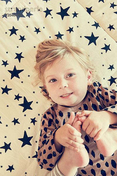 Porträt eines weiblichen Kleinkindes  das auf einem Bett mit Sternenmuster liegt und ihre nackten Füße hält
