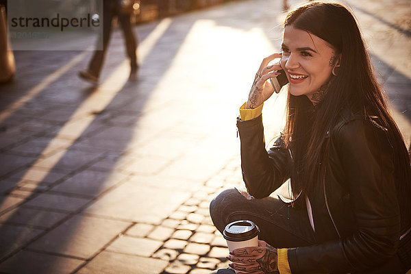 Junge Frau sitzt im Freien  hält Kaffeetasse  benutzt Smartphone  Tätowierungen an den Händen
