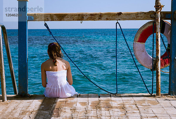 Rückansicht eines auf dem Pier sitzenden Mädchens mit Blick aufs Meer