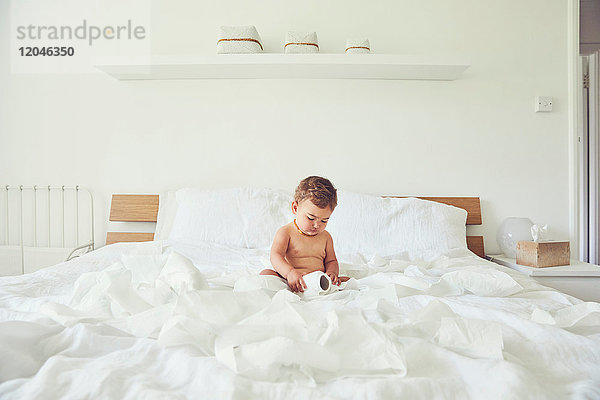 Kleinkind sitzt auf dem Bett und hält aufgetrennte Toilettenpapierrolle