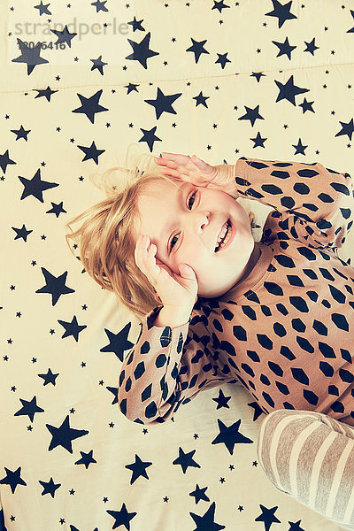 Porträt eines weiblichen Kleinkindes auf einem Sternenmusterbett liegend mit Händen auf den Wangen