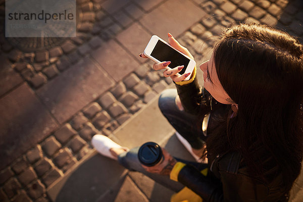 Junge Frau sitzt im Freien  hält Kaffeetasse  benutzt Smartphone  Tätowierungen auf den Händen  erhöhte Ansicht
