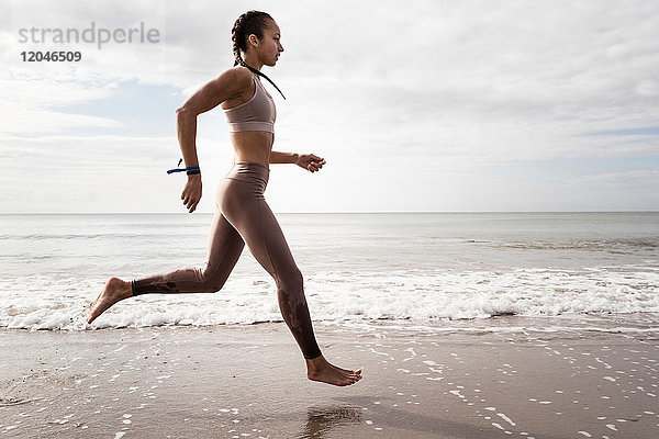 Seitenansicht einer jungen Läuferin  die am Strand barfuss am Wasser entlang läuft