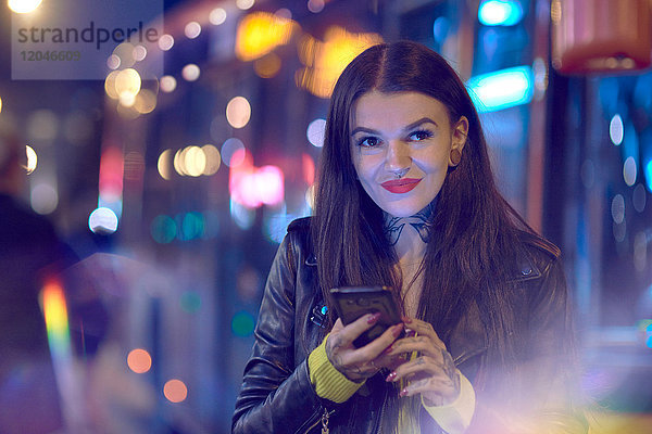 Junge Frau nachts im Freien  Smartphone in der Hand haltend  Tätowierungen an Hand und Hals