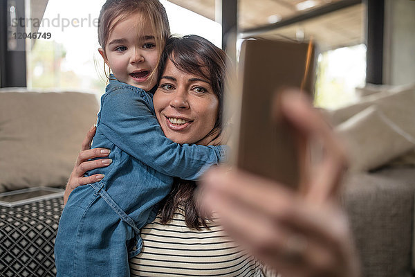 Reife Frau nimmt Smartphone-Selfie mit Tochter  die sie im Wohnzimmer umarmt