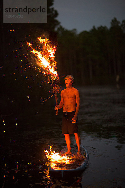 Junger Mann steht in der Dämmerung auf einem Paddelbrett auf dem Wasser  hält brennenden Ast  Flammen auf dem Paddelbrett