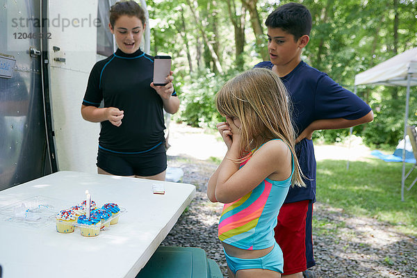 Drei Kinder stehen neben einem Tisch mit Kuchen  Kerze in Kuchen  jüngeres Mädchen schaut überrascht