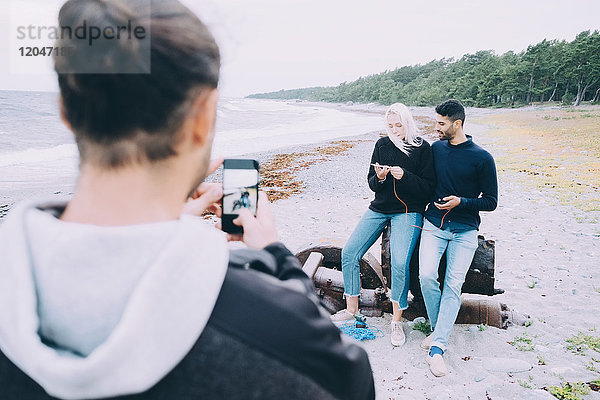 Rückansicht eines jungen Mannes  der Freunde fotografiert  die am Strand auf Metall sitzen.