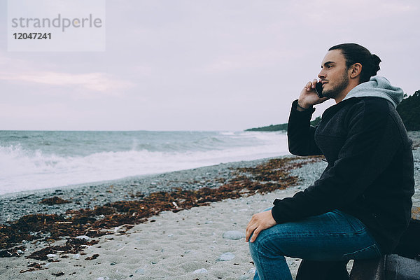 Seitenansicht eines jungen Mannes  der am Strand sitzt und sein Handy benutzt.