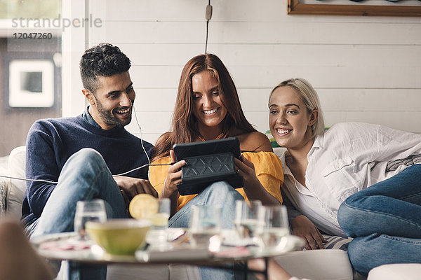 Fröhliche junge Freunde teilen sich ein digitales Tablett  während sie zu Hause auf dem Sofa sitzen.