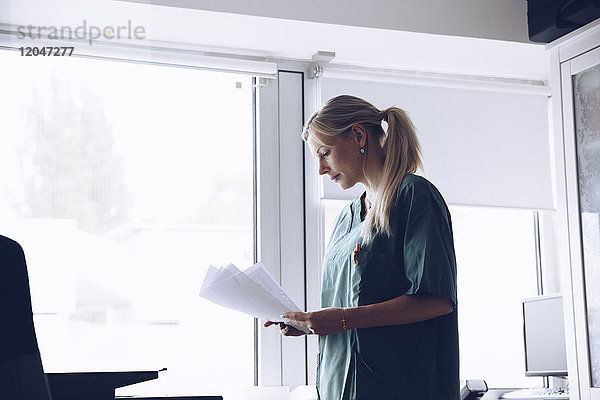 Krankenschwester liest Dokumente  während sie am Fenster des Arztes steht.