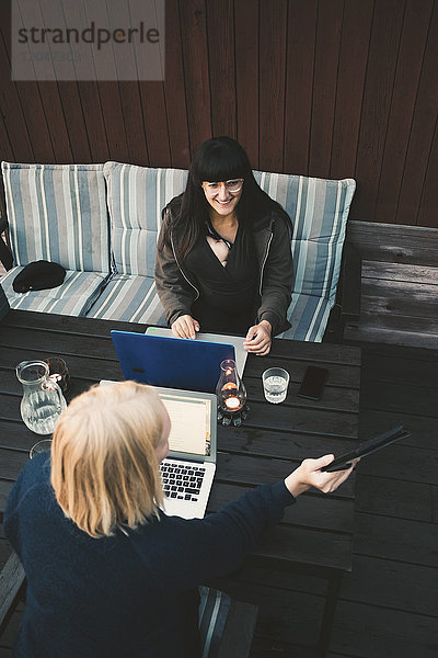 Hochwinkelansicht einer Frau  die ein digitales Tablett an eine Freundin zeigt  die einen Laptop am Tisch benutzt