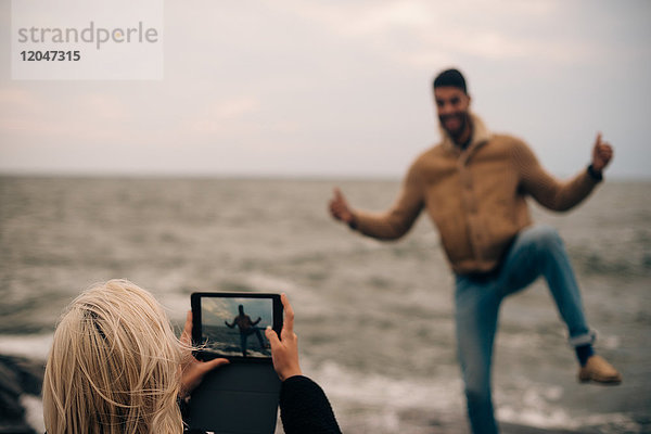 Junge Frau fotografiert Mann auf digitalem Tablett am Strand