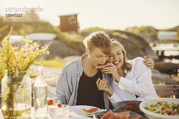 Lächelnde Frau füttert ihren Freund mit Wassermelone  während sie an einem sonnigen Tag am Picknicktisch sitzt.