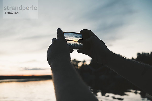 Abgeschnittenes Bild eines Mannes  der den See bei Sonnenuntergang durch ein Smartphone fotografiert.