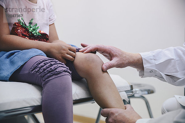 Abgeschnittene Hände eines reifen männlichen Arztes  der das Knie eines Mädchens im Krankenhaus untersucht.