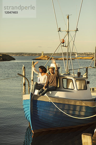 Paar mit erhobenen Händen  das im Sommer auf einem festgemachten Boot gegen den klaren Himmel sitzt.