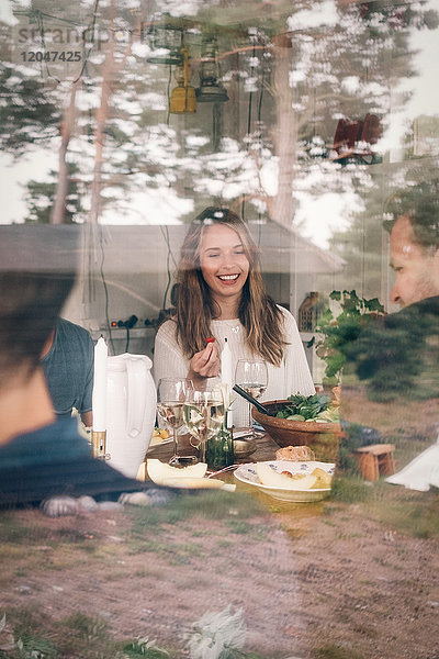 Fröhliche junge Frau beim Mittagessen mit Freunden durch Glas gesehen