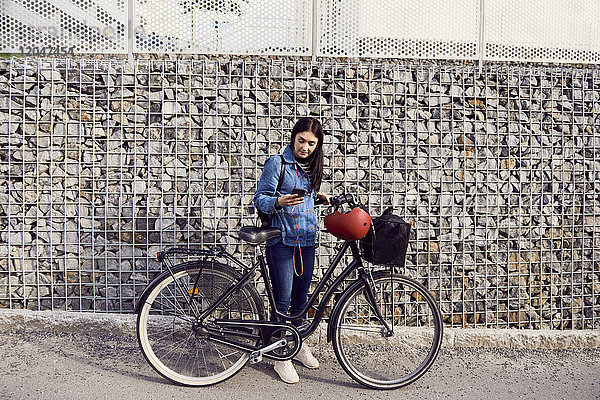 Volle Länge der Frau mit Smartphone beim Stehen mit dem Fahrrad an der Steinwand