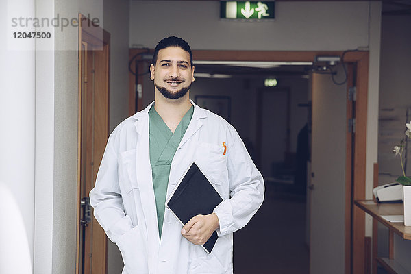 Porträt eines lächelnden jungen Arztes mit Notizblock im Krankenhausflur