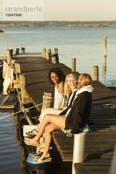 Fröhliche junge Freundinnen genießen den Sommer auf dem Steg am Hafen gegen den Himmel.