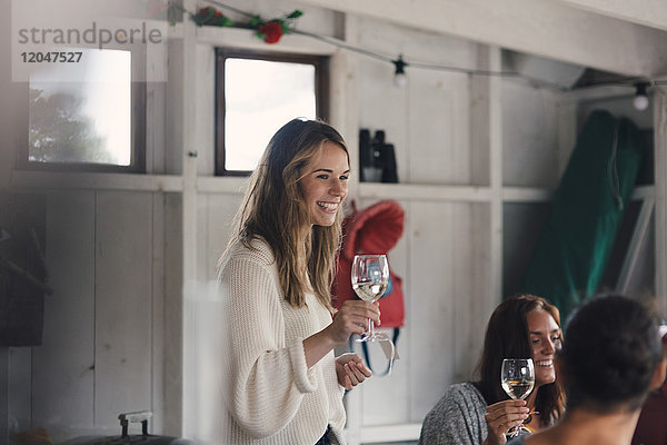 Fröhliche junge Frau steht mit Weißwein bei Freunden zu Hause
