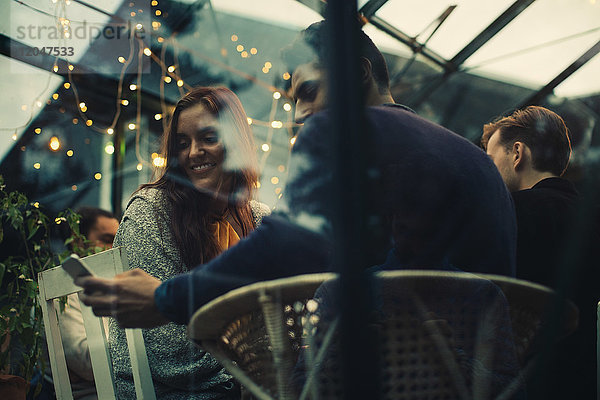 Niedriger Blickwinkel des Mannes  der dem weiblichen Freund ein Smartphone zeigt  während er während der Dinnerparty im Wintergarten sitzt.